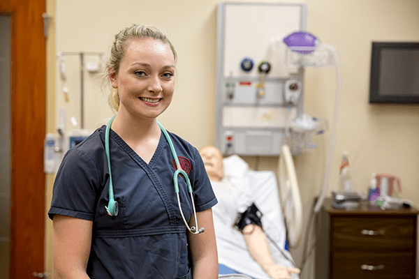 Female nursing student looking at the camera wearing gray scrubs standing in front of simulation mannequin