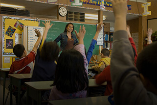 Teacher at front of classroom while students raise their hands