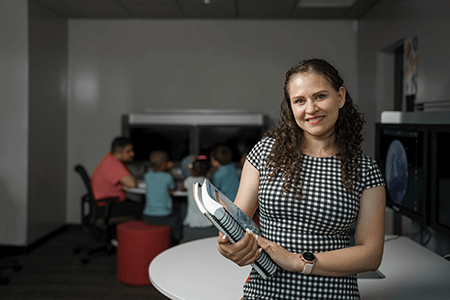 Woman looking at the camera smiling with books in her hands and children in the background learning